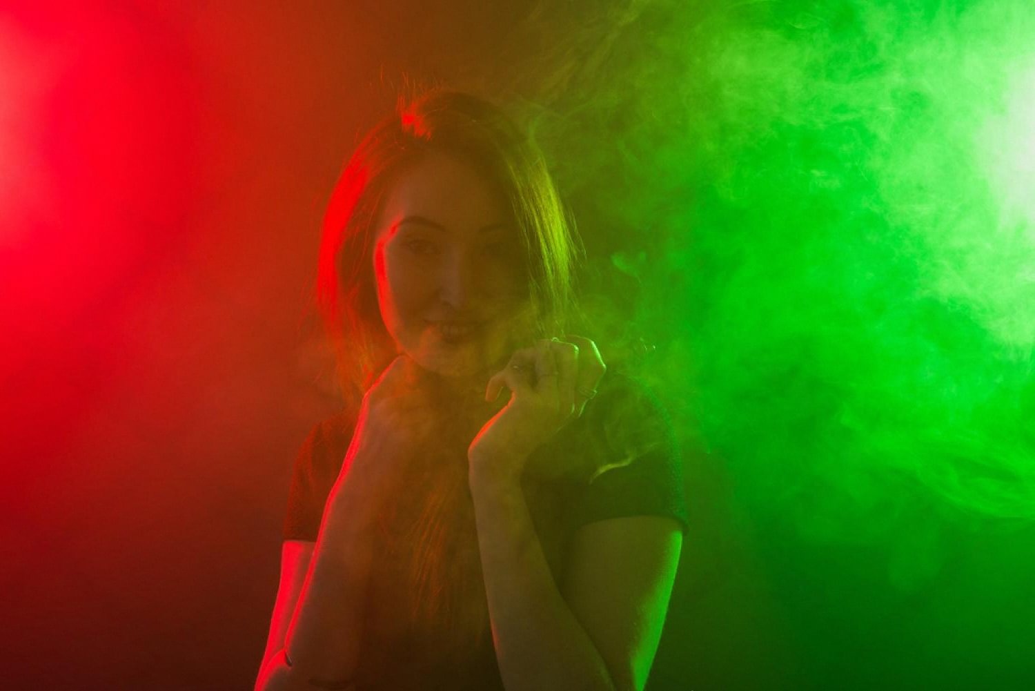 Young adult vaping a pod device under red and green studio lighting