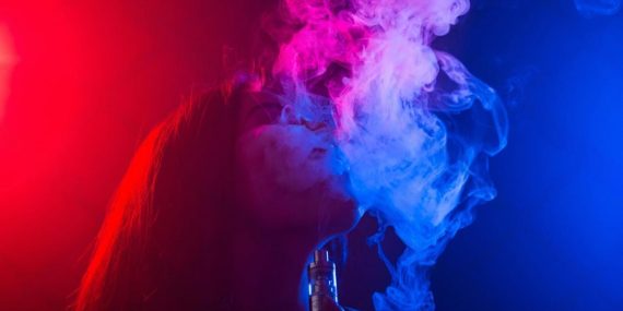 Person using a compact pod vape device with visible vapour clouds and neon lighting